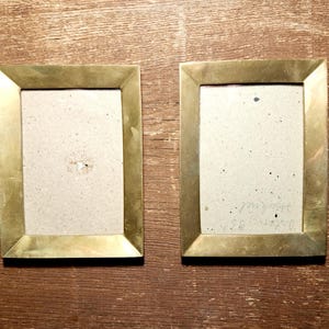 Può includere: Due cornici rettangolari in ottone con un retro marrone chiaro. Le cornici sono posizionate fianco a fianco su una superficie di legno. Le cornici hanno un design semplice e classico.