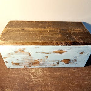 Könnte beinhalten: Eine rechteckige Holzkiste mit einem verwitterten, abgenutzten Aussehen. Der Deckel ist dunkelbraun, während der Korpus hellblau gestrichen ist, mit abblätternder Farbe, die das Holz freilegt. Die Kiste ist wahrscheinlich Vintage oder rustikal.