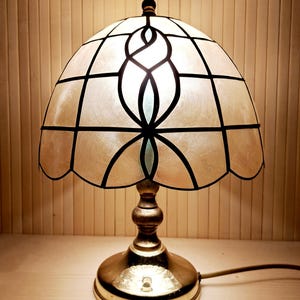 Può includere: Una lampada da tavolo decorativa con paralume in vetro colorato. Il paralume è color crema con linee nere e un disegno centrale. La base è dorata e la lampada è accesa, illuminando la stanza.
