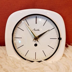 Könnte beinhalten: Ein weißer Retro-Wecker mit quadratischem Zifferblatt und abgerundeten Ecken. Das Zifferblatt ist weiß mit schwarzen Stundenmarkierungen und Zeigern. Auf dem Zifferblatt ist der Markenname "Mauthe" aufgedruckt. Die Uhr steht auf einer weißen, flauschigen Oberfläche.