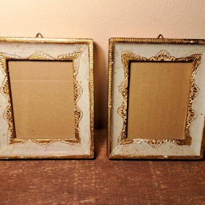 Puede incluir: Dos marcos de fotos antiguos con un acabado blanco y dorado envejecido. Los marcos son cuadrados con detalles dorados ornamentados. Cada marco tiene un pequeño gancho de metal para colgar fotos.