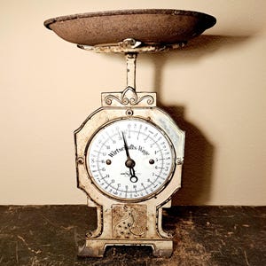 Peut inclure: Une balance mécanique vintage blanc cassé avec un cadran circulaire portant l'inscription Wirtschafts-Wage. La balance possède un plateau de pesée en métal sombre et patiné sur le dessus et un motif de paon en relief sur la base.