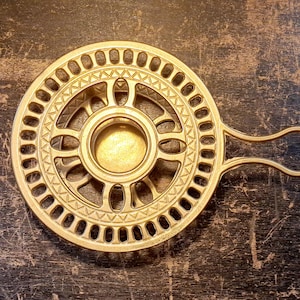 Könnte beinhalten: Ein runder Kerzenhalter aus goldfarbenem Metall mit einem zentralen Becher, einem strahlenförmigen Speichendesign und einem dekorativen Außenrand mit kleinen rechteckigen Aussparungen. Das Objekt hat einen einzelnen geschlossenen Griff.