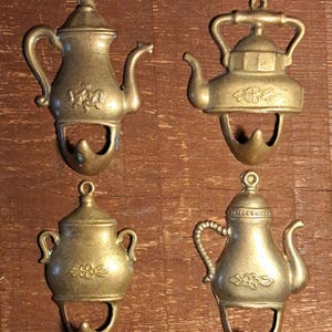 Könnte beinhalten: Vier antike, messingfarbene Wandhaken in Teekannenform. Jeder Haken hat ein Teekannen-Design mit einer Schlaufe zum Aufhängen oben und einem gebogenen Haken unten. Die Haken werden auf einer Holzoberfläche präsentiert.