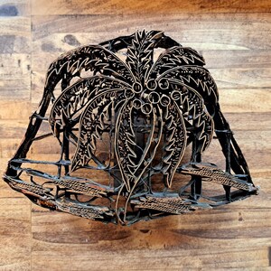 May include: A decorative, openwork metal sculpture featuring a palm tree design. The sculpture is primarily black with bronze accents, and is set against a wooden background. The palm tree has detailed fronds and coconuts.