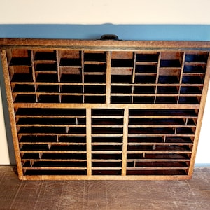 May include: A vintage wooden printer's tray with numerous small compartments. The tray is made of light brown wood and has a dark handle at the top. The interior is divided into various rectangular and square spaces, ideal for organizing small items.