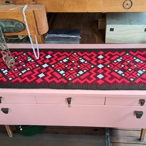 Könnte beinhalten: Ein rosa lackierter Holzschrank mit drei Schubladen, mit einem dekorativen rot, braun und weiß gemusterten Textilläufer. Eine Halskette und eine Metallkette hängen von oben. Der Schrank hat dunkle Beschläge und Holzbeine.