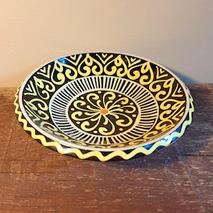Könnte beinhalten: Dekorative Keramikplatte mit einem schwarzen, gelben und weißen Design. Die Platte hat ein zentrales Blumenmuster mit radialen Linien und einem gewellten Rand. Das Farbschema umfasst Schwarz, Gelb, Weiß und einen Hauch von Orange.