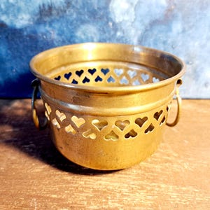 Könnte beinhalten: Ein kleiner, antiker Messing-Eimer mit herzförmigen Ausschnitten in der Mitte. Der Eimer hat zwei kleine Griffe und einen leicht ausgestellten Rand. Das Messing hat eine warme, gealterte Patina, was darauf hindeutet, dass es Vintage ist.