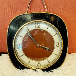 Könnte beinhalten: Vintage Wanduhr mit einem schwarzen und goldenen Rahmen und einem Griff. Das Zifferblatt ist weiß mit schwarzen Ziffern und Zeigern und einem braunen Zentrum. Die Uhr ist im Retro-Stil.