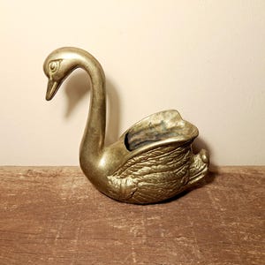 Könnte beinhalten: Eine goldfarbene Schwanfigur aus Messing mit gebogenem Hals und detailliertem Federdesign. Der Schwan hat eine kleine Öffnung auf dem Rücken. Die Figur steht auf einer Holzoberfläche. Es ist wahrscheinlich ein dekorativer Gegenstand.