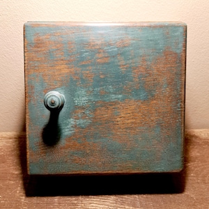 May include: A small, square wooden box with a distressed teal and brown finish. The box has a round knob on the front. The wood grain is visible through the paint, giving it a rustic appearance. The box is likely used for storage.