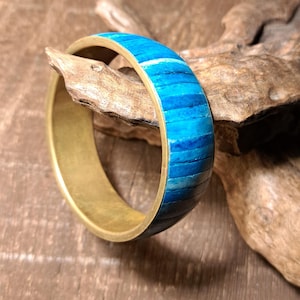 Könnte beinhalten: Ein Messingarmreif mit einem lebendigen blau-weißen Streifendesign. Das Armband hat ein hellgoldenes Band, und die blauen Streifen bestehen aus kleinen, rechteckigen Teilen. Das Armband liegt auf einem Stück Treibholz.