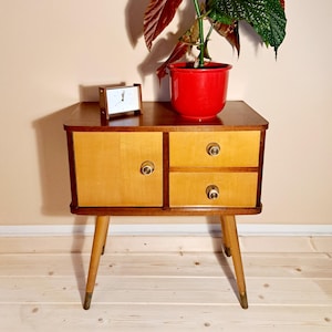 Op de afbeelding: Een vintage nachtkastje met twee lades en twee deuren, gemaakt van licht hout met een donkere rand. Een kleine klok en een rode plantenpot staan op de bovenkant. De poten zijn taps toelopend.