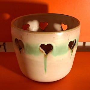 Könnte beinhalten: Eine Keramik-Tasse mit herzförmigen Aussparungen und einem grünen Band. Die Tasse hat drei kleine rote herzförmige Objekte im Inneren. Die Tasse steht auf einer orangefarbenen Oberfläche.