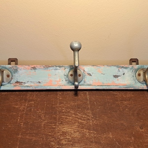 May include: A distressed wooden coat rack with three metal hooks. The wood is painted in shades of blue and pink, with a worn, vintage appearance. The hooks are silver-colored and have a rounded top.