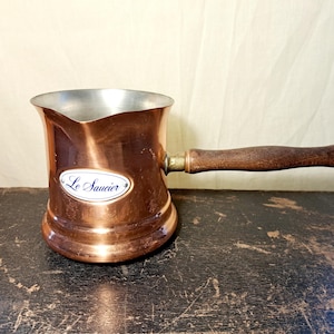 May include: A copper sauce pot with a silver-toned interior, a dark brown wooden handle, and a pouring spout. An oval white label on the front features the text Le Saucier in blue script. The pot has a wide base and a polished metallic finish.