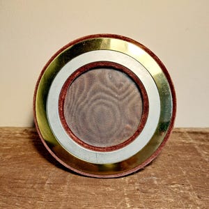 Könnte beinhalten: Ein runder Bilderrahmen mit bordeauxrotem Samtrücken. Der Rahmen hat einen goldfarbenen Außenring, einen silberfarbenen Innenring und eine mit Mesh bedeckte Mitte. Er steht auf einer Holzoberfläche.