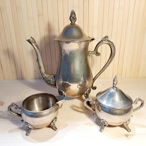 Könnte beinhalten: Ein silbernes Teeservice mit aufwendigen Details. Das Set besteht aus einer Teekanne, einem Sahnekännchen und einer Zuckerdose. Die Teekanne hat einen gebogenen Ausguss und einen dekorativen Griff. Sahnekännchen und Zuckerdose haben passende Griffe und stehen auf kleinen Füßen.