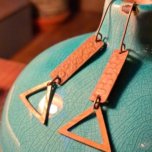 May include: A pair of copper-coloured earrings. Each earring has a rectangular top with a textured surface and a triangle-shaped pendant. The earrings hang from a simple wire hook. The background is a turquoise ceramic vase.