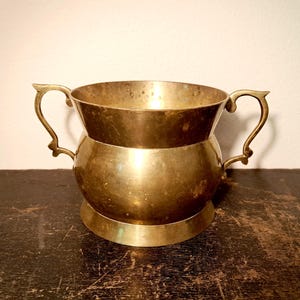 Puede incluir: Un cuenco de latón vintage con dos asas ornamentadas. El cuenco tiene un borde ancho y acampanado y un cuerpo redondeado, que se asienta sobre una base circular. El latón tiene un tono dorado cálido y muestra signos de edad y uso.