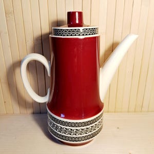 Könnte beinhalten: Eine rot-weiße Keramik-Kaffeekanne mit Deckel und Griff. Die Kanne hat einen weißen Ausguss und Griff sowie ein schwarz-weiß gemustertes Band um den Boden und den Deckel. Der Hintergrund ist helles Holz.