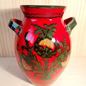 May include: A vibrant red ceramic vase with a glossy finish. The vase features a floral design in green and gold, with two handles. The rim and handles are accented with dark green. This decorative vase is suitable for home decor.