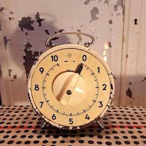 May include: A vintage cream-colored kitchen timer with black numbers and a central dial. The timer has a metal handle and feet, showing signs of wear. The dial is set to approximately 1 minute. The background is a distressed white surface.