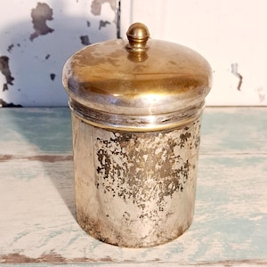 Könnte beinhalten: Ein silberfarbenes Vintage-Glas mit einem goldfarbenen Deckel und einem kleinen Knopf. Das Glas hat eine zylindrische Form mit einer strukturierten, abgenutzten Oberfläche. Der Deckel ist abgerundet und glänzend, im Gegensatz zum gealterten Aussehen des Glaskörpers. Das Glas steht auf einer hellblau-weißen Oberfläche.