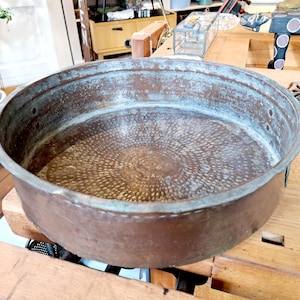 Può includere: Un grande bacino antico in rame con un bordo largo e un interno strutturato e dettagliato. Il bacino ha un aspetto invecchiato e usurato con patina visibile e due piccole maniglie sui lati. L'interno presenta un motivo circolare a pois.