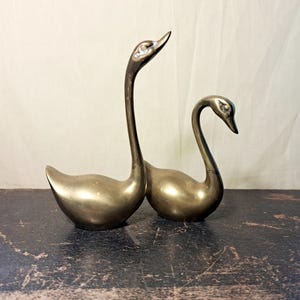 Könnte beinhalten: Zwei Schwanenfiguren aus Messing mit polierter Metalloberfläche. Der größere Schwan hat einen langen, nach oben gerichteten Hals, während der kleinere Schwan einen kürzeren, nach unten gebogenen Hals hat. Beide Figuren haben glatte Körper.
