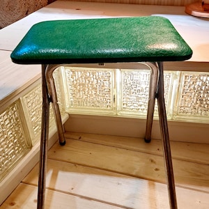Pode incluir: Banqueta vintage com assento retangular revestido em vinil verde. A banqueta tem uma estrutura de metal cromado e quatro pernas anguladas. O assento tem aproximadamente 30 cm de largura e 25 cm de profundidade.