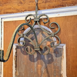 Könnte beinhalten: Ein verzierter, grüner Metall-Hängehalter mit Schnörkeln und einem gebogenen Arm. Der Halter ist an einem Haken aufgehängt und an einer Holztür mit weißer Zierleiste befestigt. Das Holz hat eine natürliche Maserung und ein verwittertes Aussehen.