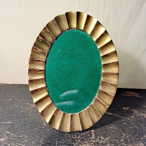 Könnte beinhalten: Ein ovaler Bilderrahmen mit einem polierten Messingrand und einem ausgeprägten geriffelten oder gewellten Design. Die Mitte des Rahmens ist mit einem einfarbigen, leuchtend grünen, filzartigen Material gefüllt, was einen kontrastreichen Effekt erzeugt.