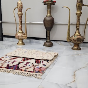 May include: An assortment of decorative items, including two ornate brass pitchers with curved spouts and a central vase. A small, colorful rug with a geometric pattern is in the foreground. The items are arranged on a tiled floor.