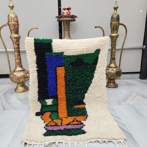 May include: A rectangular, off-white rug with a colorful abstract design. The design features blocks of black, blue, green, orange, and purple. The rug has a fringe along the bottom edge. Decorative brass pitchers flank the rug.