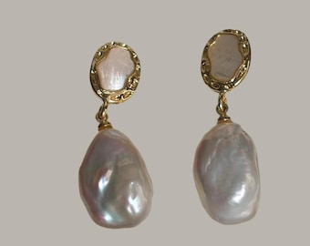 Pode incluir: Um par de brincos pendurados dourados. Cada brinco apresenta uma pequena parte superior ovalada esbranquiçada com uma borda dourada decorativa, conectada a uma grande pérola iridescente em forma de gota. As pérolas têm um brilho perolado.