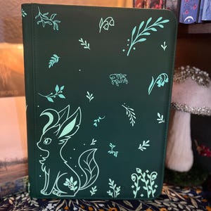 May include: A dark green rectangular notebook cover featuring a light mint green graphic of a stylized fox-like creature with large ears and a crescent moon shape on its head, surrounded by various small botanical leaf and flower illustrations.