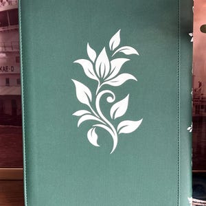 May include: A rectangular sage green notebook with a textured cover featuring a centered white floral graphic. The design consists of a stylized flower with leaves and a curved stem. The notebook has rounded corners and a vertical spine detail.
