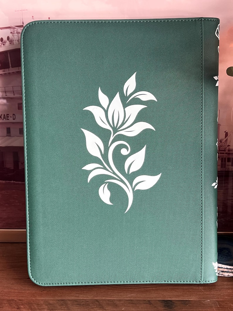 May include: A rectangular sage green notebook with a textured cover featuring a centered white floral graphic. The design consists of a stylized flower with leaves and a curved stem. The notebook has rounded corners and a vertical spine detail.