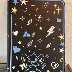 May include: A black rectangular binder featuring a repeating pattern of light blue, yellow, and white stars, lightning bolts, and hearts. A cartoon character resembling a small creature with large ears is centered at the bottom.