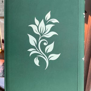 May include: A rectangular dark green binder with a textured surface featuring a centered white floral graphic. The design consists of a stylized flower with multiple leaves and a curved stem. The spine of the binder is visible on the right side.