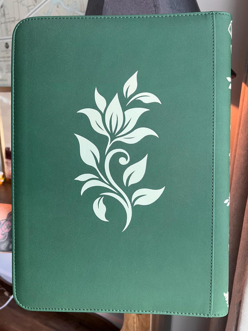 May include: A rectangular dark green binder with a textured surface featuring a centered white floral graphic. The design consists of a stylized flower with multiple leaves and a curved stem. The spine of the binder is visible on the right side.