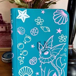 May include: A teal binder featuring a repeating pattern of white and pink line art illustrations. The design includes a central Vaporeon character, various seashells, starfish, and bubbles scattered across the front cover surface.