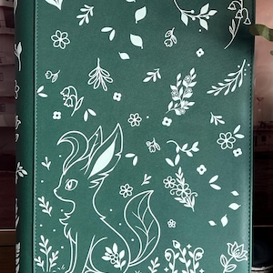 May include: A dark green rectangular binder featuring a white line art illustration of a fox-like creature sitting among various scattered floral and leaf motifs. The cover has a textured finish and a vertical spine detail on the left side.