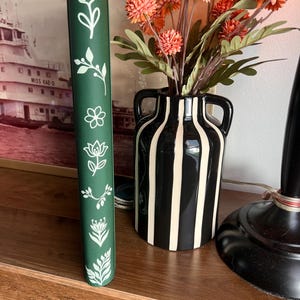 May include: A dark green vertical planner or notebook spine featuring a series of white line-art botanical illustrations including various flowers, leaves, and sprigs arranged in a single column along the center of the textured cover surface.