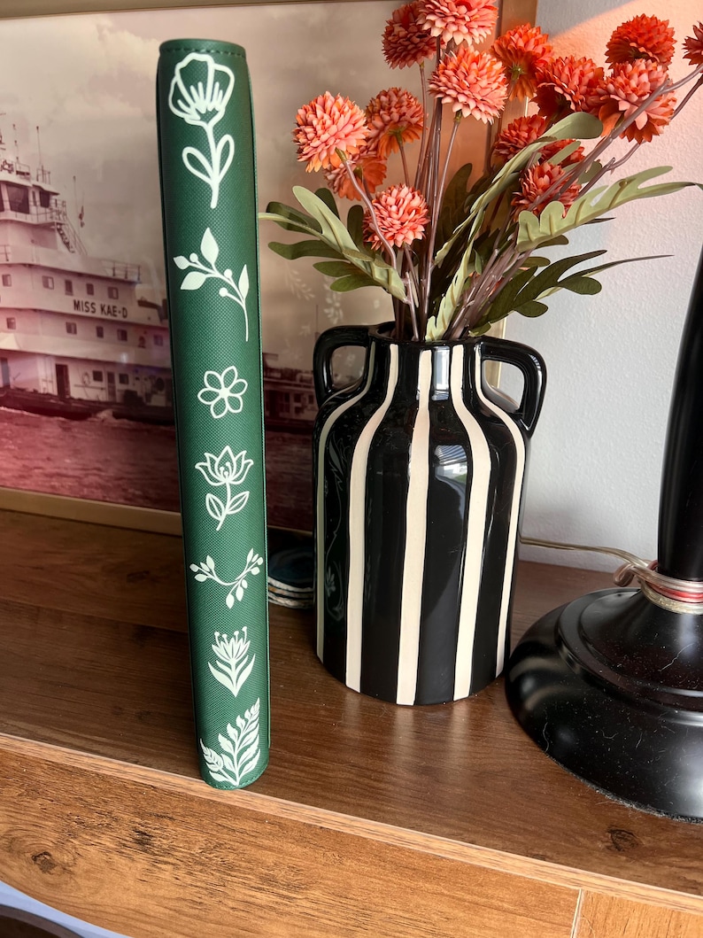 May include: A dark green vertical planner or notebook spine featuring a series of white line-art botanical illustrations including various flowers, leaves, and sprigs arranged in a single column along the center of the textured cover surface.