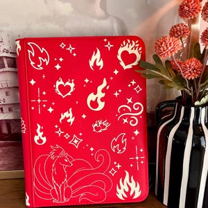 May include: A bright red rectangular binder featuring a white line art pattern of various flame shapes, hearts, and sparkling stars. At the bottom, a large illustration of a multi-tailed fox is depicted in white lines against the red background.