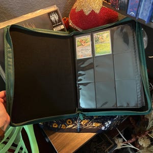 May include: A dark green zippered trading card binder featuring a black interior lining. The right side contains clear plastic sleeves with two visible trading cards inserted into the top row, while the remaining slots are empty.