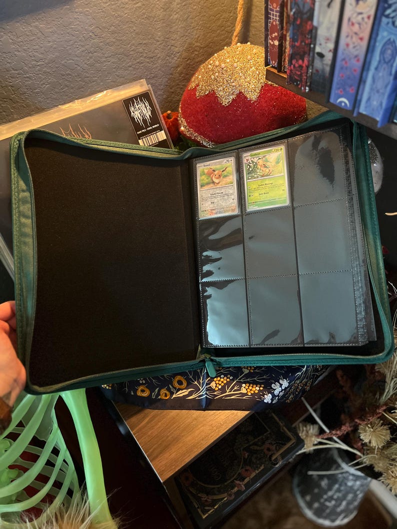 May include: A dark green zippered trading card binder featuring a black interior lining. The right side contains clear plastic sleeves with two visible trading cards inserted into the top row, while the remaining slots are empty.